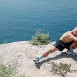 Shirtless man doing a stretch on a cliff overlooking the ocean in daylight.