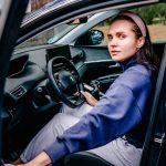 Woman sitting in the driver's seat of a car with an open door, looking outside. Natural setting.